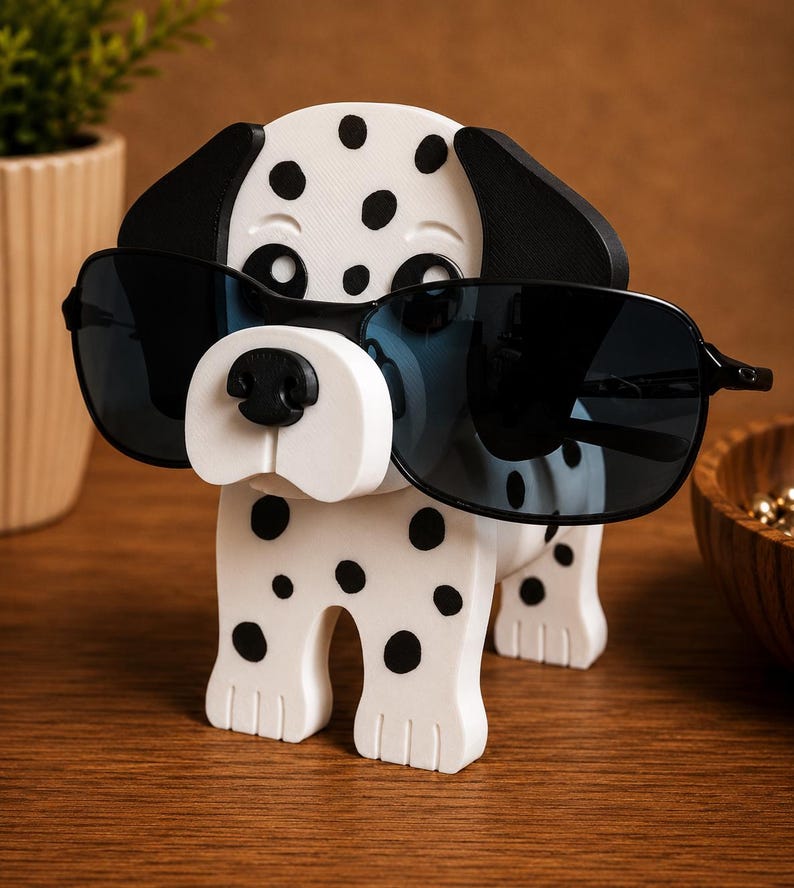 K&ouml;nnte beinhalten: Ein wei&szlig;-schwarzer Dalmatiner-Sonnenbrillenhalter. Der Halter hat schwarze Sonnenbrillen auf der Nase. Der Hund hat schwarze Ohren und schwarze Flecken auf seinem K&ouml;rper. Der Halter steht auf einer Holzoberfl&auml;che.