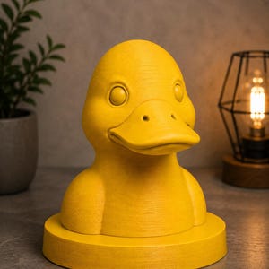 May include: A bright yellow duck-shaped decorative object. The duck has a detailed beak, eyes, and a textured surface. It sits on a round base. The background includes a plant in a pot and a lamp.