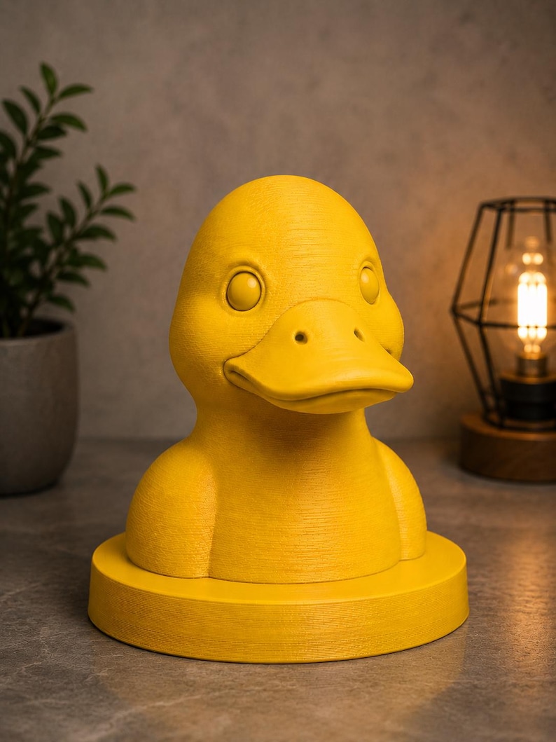 May include: A bright yellow duck-shaped decorative object. The duck has a detailed beak, eyes, and a textured surface. It sits on a round base. The background includes a plant in a pot and a lamp.