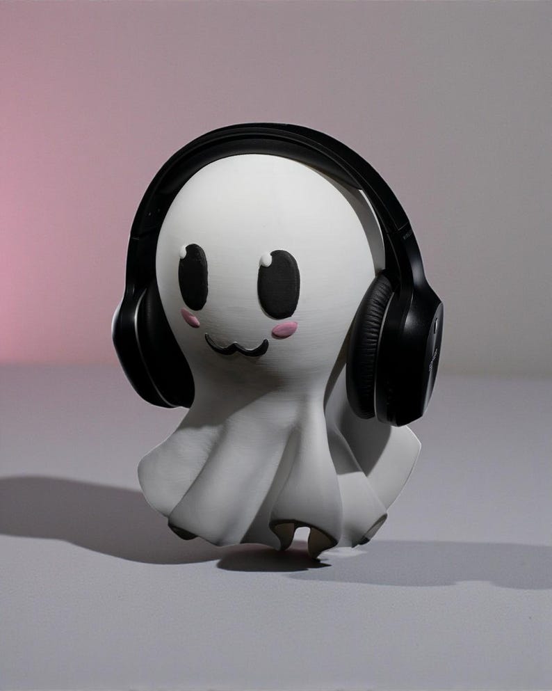 May include: A white ghost-shaped figurine with black eyes, a small black mouth, and pink cheeks, wearing black headphones. The figurine has a draped, flowing design, and is set against a gradient pink and gray background.