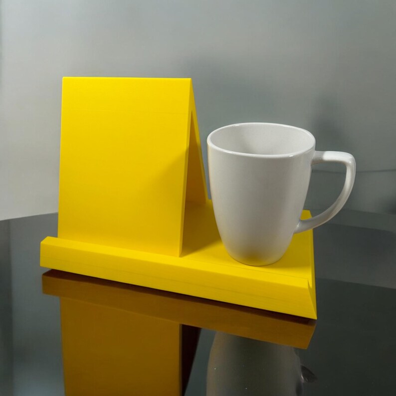 May include: A bright yellow, angled display stand with a white ceramic mug. The stand has a flat base and a vertical back panel. The mug has a curved handle and a smooth, glossy finish. The mug is on the right side of the stand.