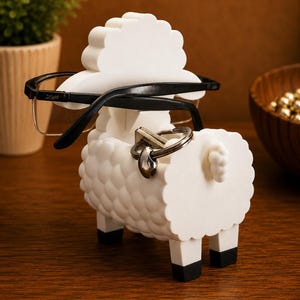 May include: A white sheep-shaped eyeglasses and key holder. The holder has a textured body, black legs, and a fluffy head. Black eyeglasses rest on the head, and a key ring hangs from the back. The holder is on a wooden surface.