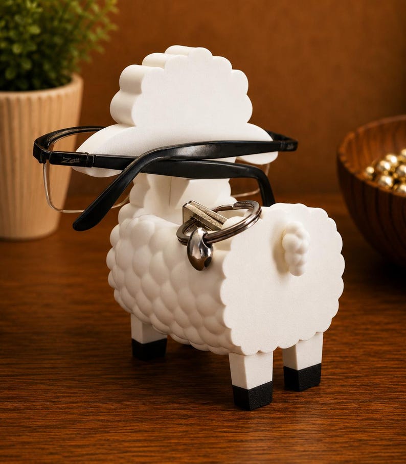 May include: A white sheep-shaped eyeglasses and key holder. The holder has a textured body, black legs, and a fluffy head. Black eyeglasses rest on the head, and a key ring hangs from the back. The holder is on a wooden surface.