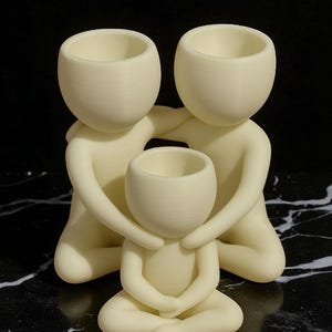 Puede incluir: Una escultura familiar de color crema. Dos figuras abrazan a una figura m&aacute;s peque&ntilde;a en el centro. Cada figura tiene una cabeza redondeada con una parte superior ahuecada, y la figura central est&aacute; sentada con las piernas cruzadas. La escultura est&aacute; sobre una superficie veteada en blanco y negro.
