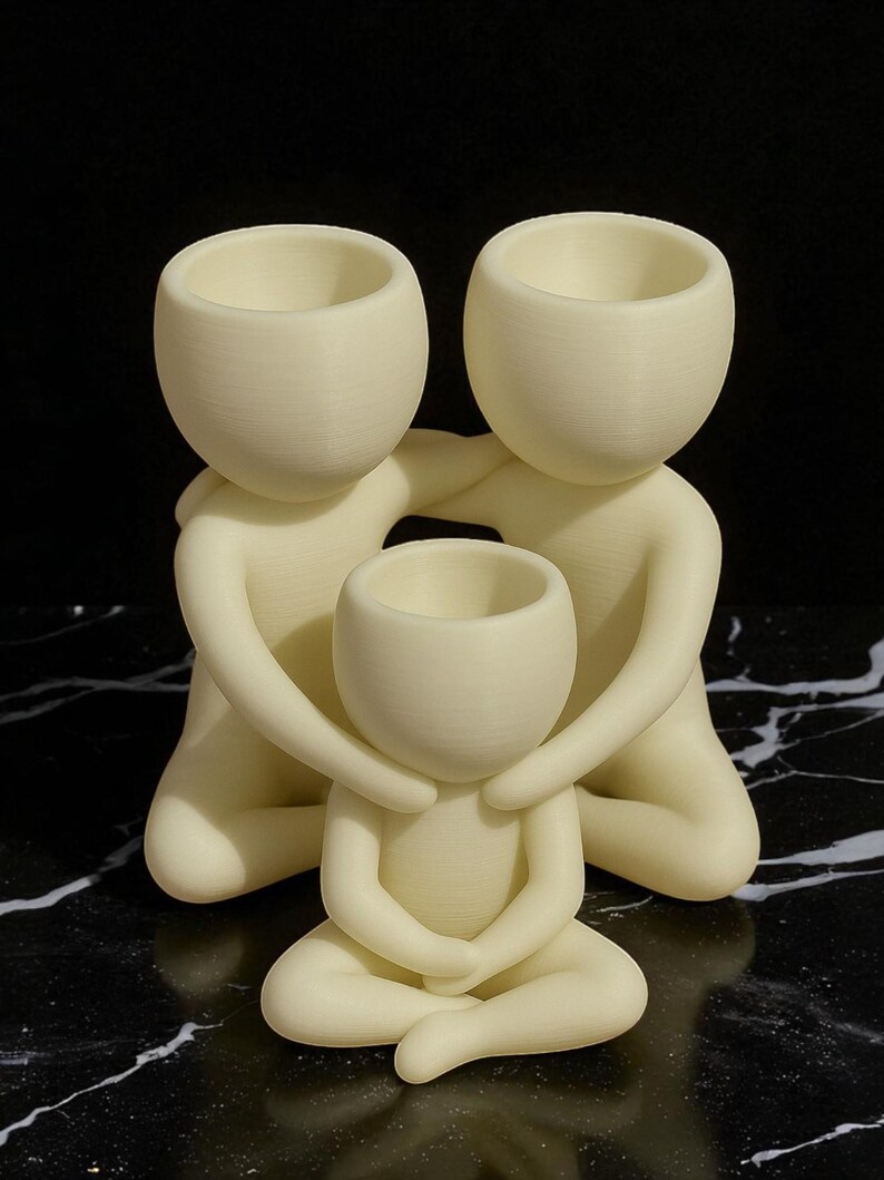 Puede incluir: Una escultura familiar de color crema. Dos figuras abrazan a una figura m&aacute;s peque&ntilde;a en el centro. Cada figura tiene una cabeza redondeada con una parte superior ahuecada, y la figura central est&aacute; sentada con las piernas cruzadas. La escultura est&aacute; sobre una superficie veteada en blanco y negro.