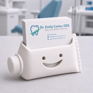 Pu&ograve; includere: Un portacarte bianco a forma di tubetto di dentifricio sorridente, che contiene una pila di biglietti da visita. Le carte presentano il testo "Dr. Emily Carter, DDS" e le informazioni di contatto. Il supporto ha un tappo e un design strutturato.