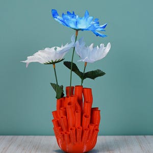 May include: An orange, coral-shaped vase holds three artificial flowers. The flowers are white and blue, with green stems and leaves. The vase is made of a solid, bright orange material and sits on a light-colored surface against a teal background.