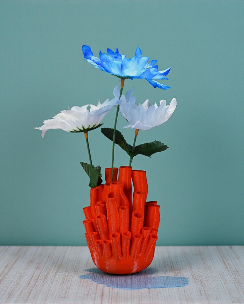 May include: An orange, coral-shaped vase holds three artificial flowers. The flowers are white and blue, with green stems and leaves. The vase is made of a solid, bright orange material and sits on a light-colored surface against a teal background.