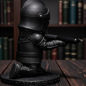 May include: A black knight figurine kneeling and holding a pen. The knight is wearing full armor, including a helmet, shoulder pads, and leg armor. The figurine is on a round base with a decorative border. The background features a bookshelf with books.