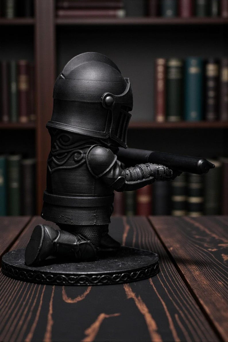 May include: A black knight figurine kneeling and holding a pen. The knight is wearing full armor, including a helmet, shoulder pads, and leg armor. The figurine is on a round base with a decorative border. The background features a bookshelf with books.