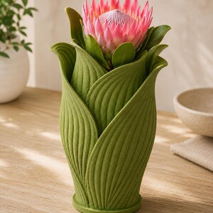 May include: A green, leaf-shaped vase holding a pink protea flower with green leaves. The vase has a textured surface and sits on a wooden table. A white bowl and a plant in a white pot are in the background.