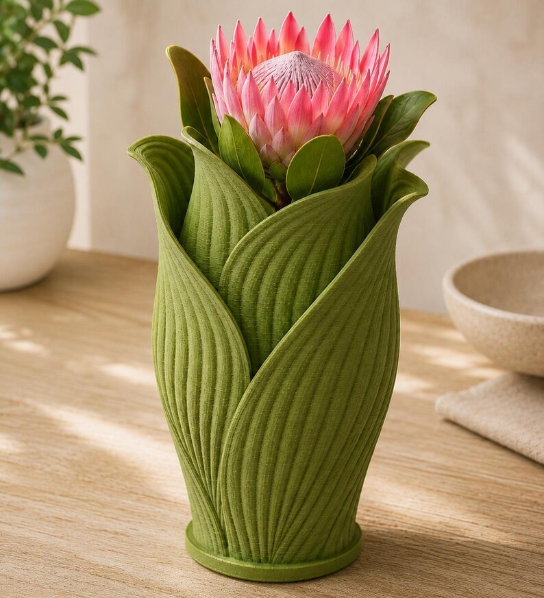 May include: A green, leaf-shaped vase holding a pink protea flower with green leaves. The vase has a textured surface and sits on a wooden table. A white bowl and a plant in a white pot are in the background.