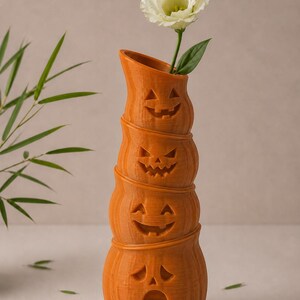 May include: Orange pumpkin-shaped vase with carved faces, stacked to create a tall, decorative piece. A white flower with green leaves is placed in the top opening. The vase is a festive Halloween decoration.
