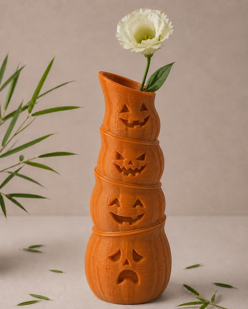 May include: Orange pumpkin-shaped vase with carved faces, stacked to create a tall, decorative piece. A white flower with green leaves is placed in the top opening. The vase is a festive Halloween decoration.