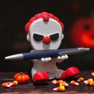 May include: A white clown figurine with red hair, nose, and feet, holding a blue pen. The clown has black eye makeup and is surrounded by candy corn and a small orange pumpkin. Halloween-themed decor.