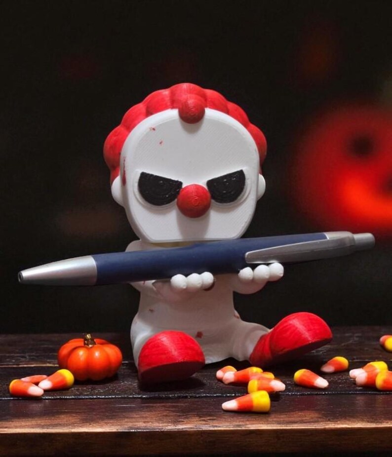 May include: A white clown figurine with red hair, nose, and feet, holding a blue pen. The clown has black eye makeup and is surrounded by candy corn and a small orange pumpkin. Halloween-themed decor.