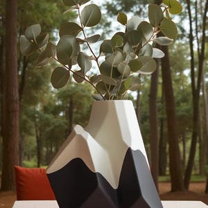 Op de afbeelding: Een moderne, geometrische vaas in zwart, wit en beige, gevormd als een bergketen. De vaas bevat een takje eucalyptus met ronde, groene bladeren. De vaas staat op een lichtgekleurde tafel met een bosachtergrond.