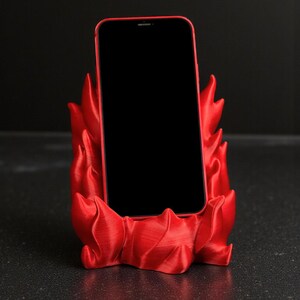 Puede incluir: Un tel&eacute;fono inteligente rojo descansa en un soporte para tel&eacute;fono rojo a juego. El soporte tiene un dise&ntilde;o en forma de llama, con bordes curvos y puntiagudos. La pantalla del tel&eacute;fono es negra, contrastando con el rojo vibrante del soporte y el marco del tel&eacute;fono.