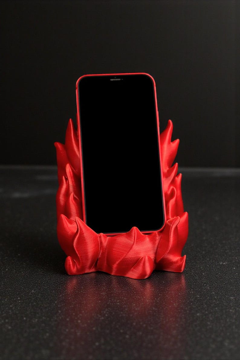Puede incluir: Un tel&eacute;fono inteligente rojo descansa en un soporte para tel&eacute;fono rojo a juego. El soporte tiene un dise&ntilde;o en forma de llama, con bordes curvos y puntiagudos. La pantalla del tel&eacute;fono es negra, contrastando con el rojo vibrante del soporte y el marco del tel&eacute;fono.