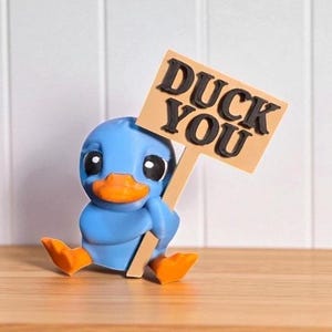 May include: A blue duck figurine with an orange beak and feet, holding a sign that reads "DUCK YOU" in black letters. The duck is sitting on a wooden surface against a white background.