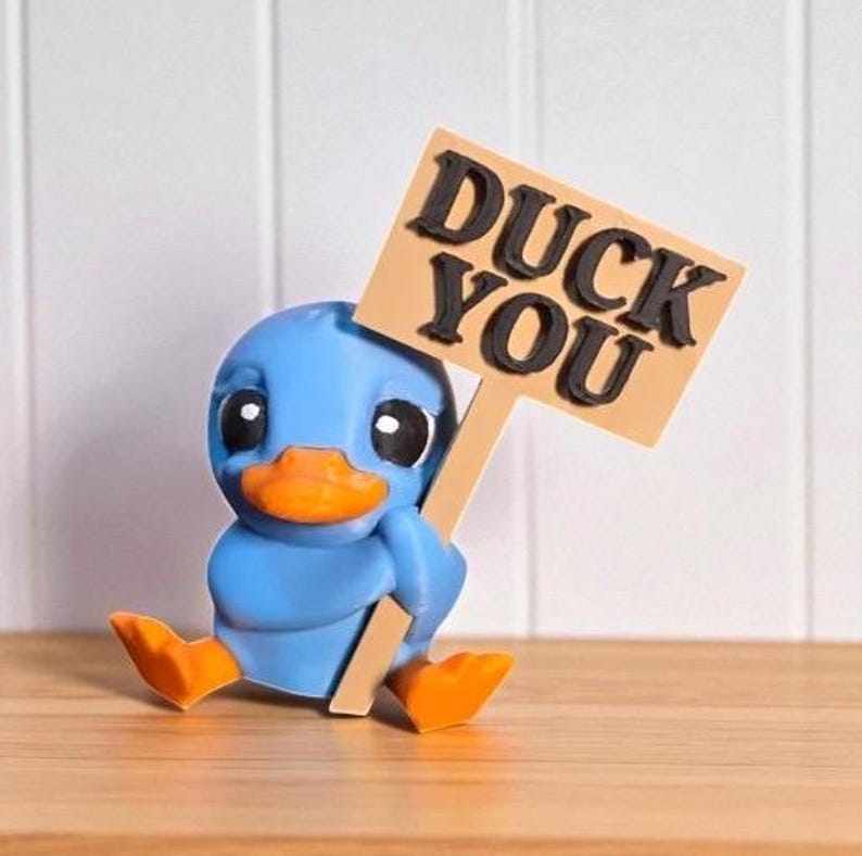 May include: A blue duck figurine with an orange beak and feet, holding a sign that reads "DUCK YOU" in black letters. The duck is sitting on a wooden surface against a white background.