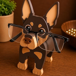 May include: A wooden dog-shaped eyeglasses holder in brown and tan. The holder has a dog's face and body, with the glasses resting on the nose. The dog has large ears and paws. The glasses have black frames.