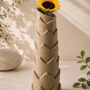 May include: A tall, beige vase with a stacked design, holding a vibrant yellow sunflower. The vase has a textured, corrugated appearance with triangular accents. The sunflower's dark center contrasts with its bright yellow petals.