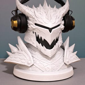 Può includere: Busto bianco a tema drago con superficie strutturata e dettagliata, con cuffie nere e dorate. Il busto presenta corna, una corona appuntita e un sorriso minaccioso. Le cuffie suggeriscono audio o gaming.