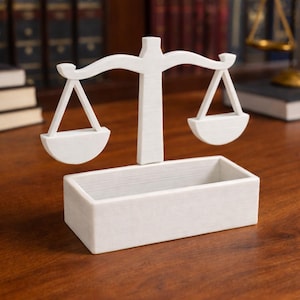 Puede incluir: Una balanza de la justicia decorativa blanca, con una base rectangular y una balanza con dos platillos colgantes. La balanza se encuentra sobre un fondo borroso de libros y una superficie de madera.