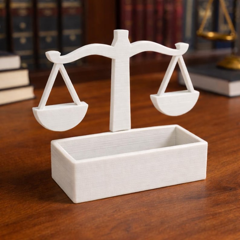 Puede incluir: Una balanza de la justicia decorativa blanca, con una base rectangular y una balanza con dos platillos colgantes. La balanza se encuentra sobre un fondo borroso de libros y una superficie de madera.