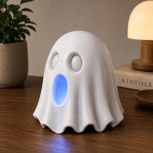 May include: A white ghost-shaped night light with a blue glow emanating from its mouth. The light has two round eye-shaped openings and a draped, sheet-like form. It sits on a wooden surface, suggesting a decorative or functional item.