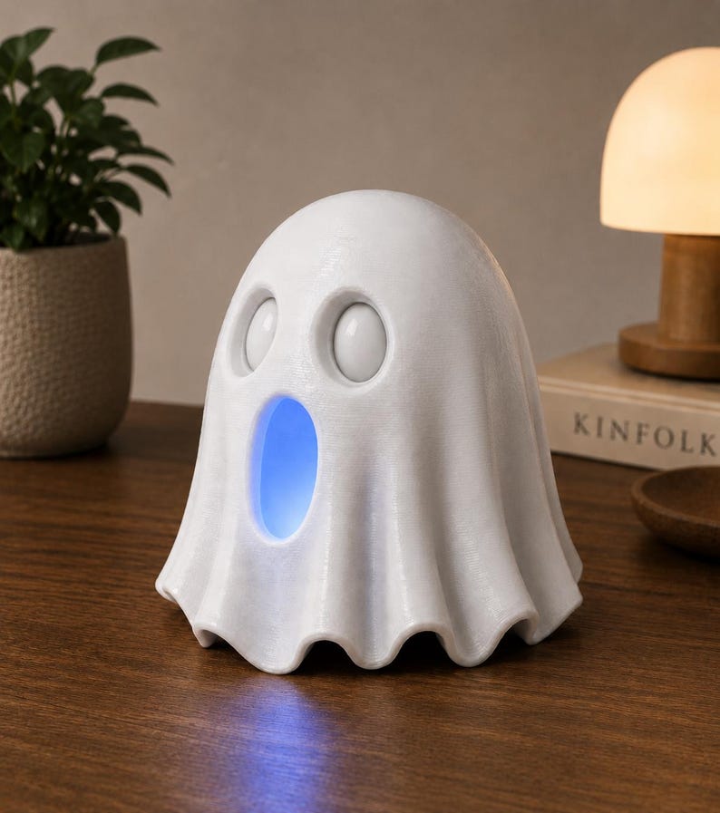 May include: A white ghost-shaped night light with a blue glow emanating from its mouth. The light has two round eye-shaped openings and a draped, sheet-like form. It sits on a wooden surface, suggesting a decorative or functional item.
