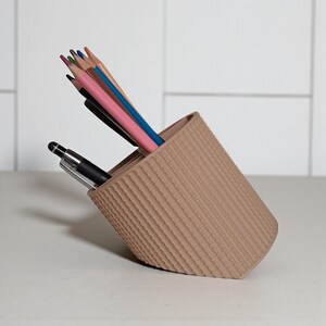 May include: A beige, textured pencil holder tilted to the side, containing several colored pencils and a black pen. The pencils include shades of pink, blue, and green. The holder has a ribbed design.
