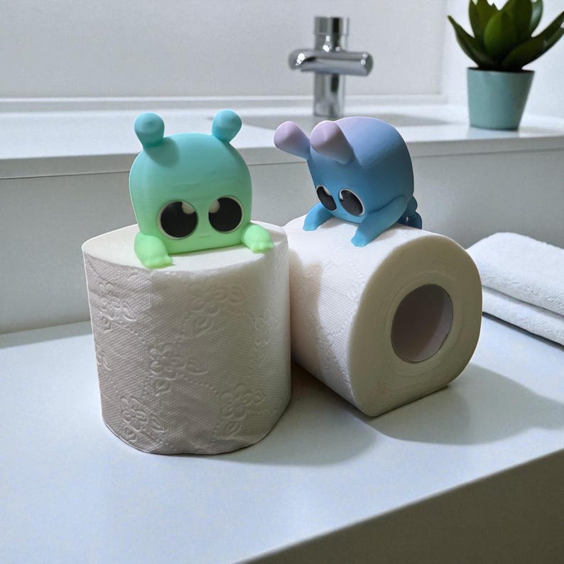 May include: Two rolls of white toilet paper are topped with whimsical, colorful, cartoon-like creatures. One is teal, the other blue, each with large black eyes and small arms. The scene is set in a bathroom.