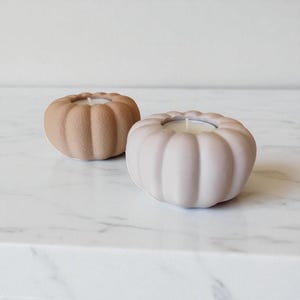 Puede incluir: Dos velas con forma de calabaza en tonos neutros. Una es de color beige claro y la otra malva p&aacute;lido. Cada vela tiene un centro de cera blanca. Las velas est&aacute;n colocadas sobre una superficie de m&aacute;rmol blanco.