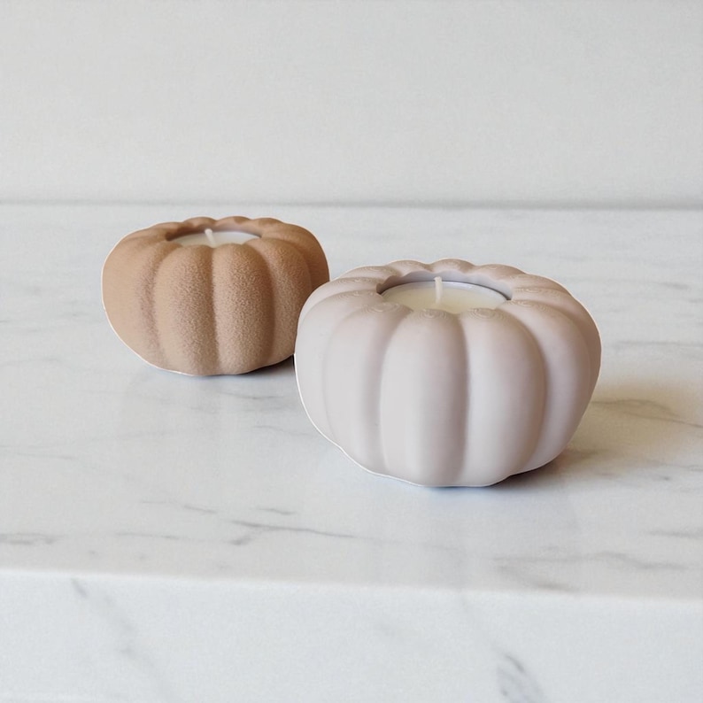 Puede incluir: Dos velas con forma de calabaza en tonos neutros. Una es de color beige claro y la otra malva p&aacute;lido. Cada vela tiene un centro de cera blanca. Las velas est&aacute;n colocadas sobre una superficie de m&aacute;rmol blanco.