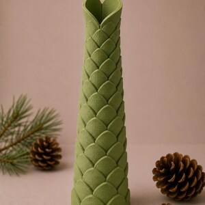 May include: A tall, green vase with a textured, scale-like pattern. The top of the vase is shaped like a heart. The vase is surrounded by pine cones and a sprig of pine needles, suggesting a nature-inspired theme.