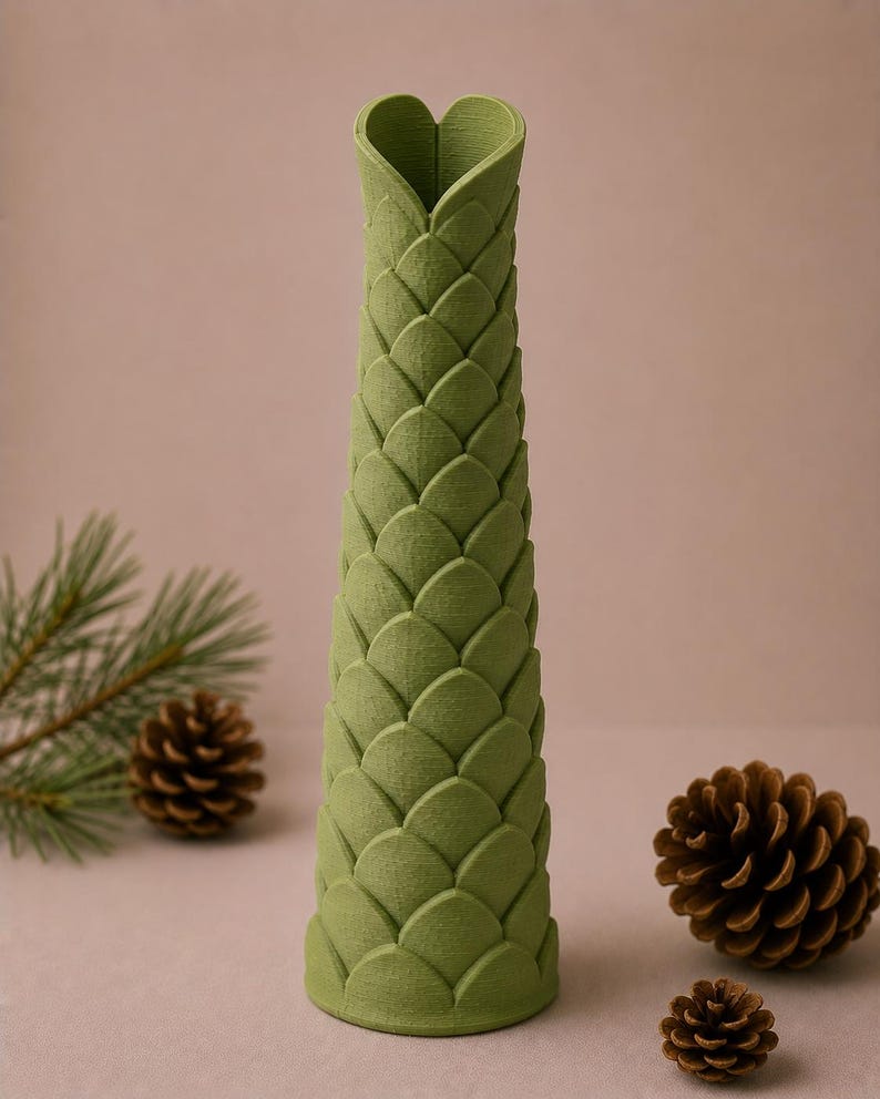 May include: A tall, green vase with a textured, scale-like pattern. The top of the vase is shaped like a heart. The vase is surrounded by pine cones and a sprig of pine needles, suggesting a nature-inspired theme.