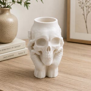 May include: A white ceramic skull-shaped vase. The skull's eye sockets and nose are hollow, and the hands are positioned as if holding the skull. The vase is a decorative item, suitable for Halloween or gothic decor.