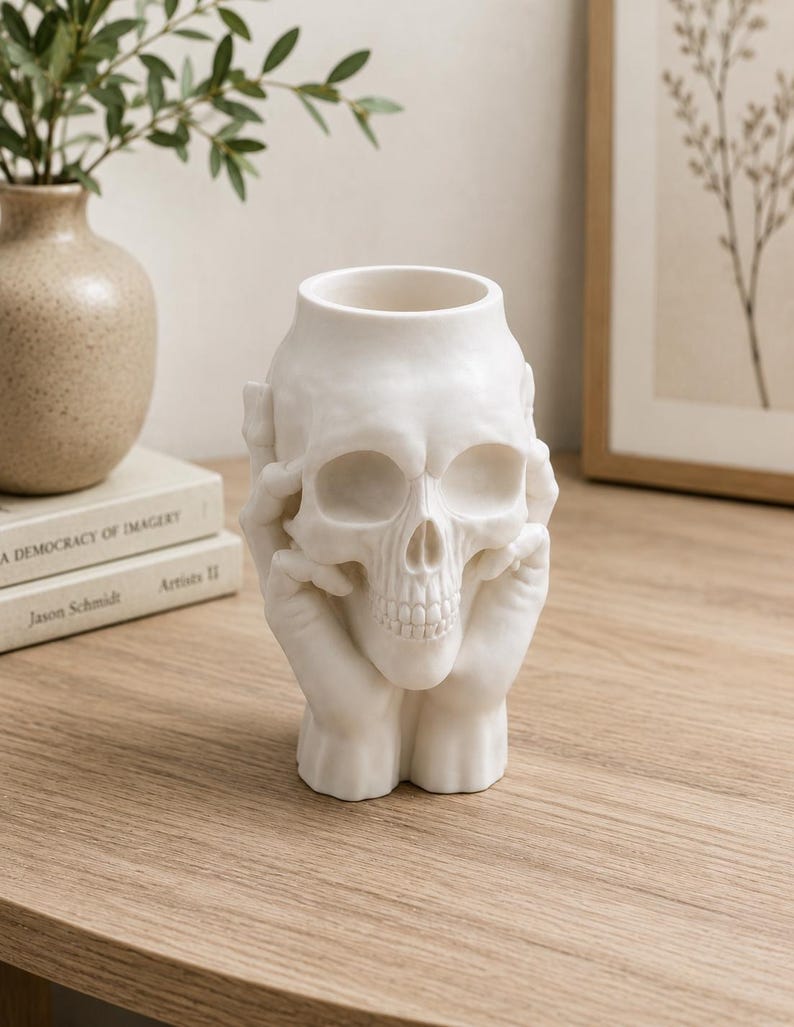 May include: A white ceramic skull-shaped vase. The skull's eye sockets and nose are hollow, and the hands are positioned as if holding the skull. The vase is a decorative item, suitable for Halloween or gothic decor.