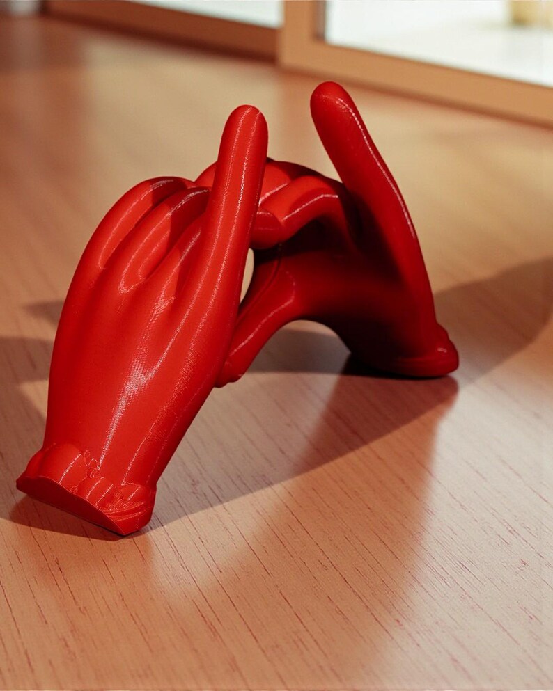 May include: A vibrant red sculpture of two hands intertwined, forming a heart shape. The hands are detailed, with visible fingers and a smooth, glossy finish. The sculpture is placed on a light-colored wooden surface, creating a striking contrast.