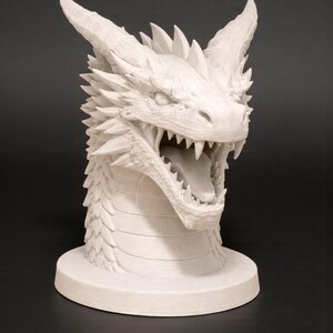 May include: A white dragon head sculpture with an open mouth, revealing sharp teeth and a long tongue. The sculpture has detailed scales, horns, and a spiky mane. The dragon head is mounted on a round base.