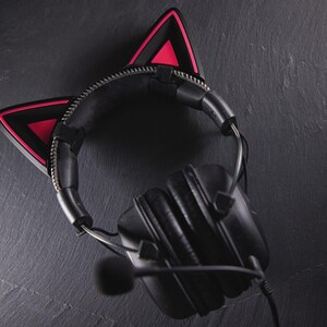 May include: Black headphones with a microphone and pink cat ear covers. The headphones are on a dark grey surface.