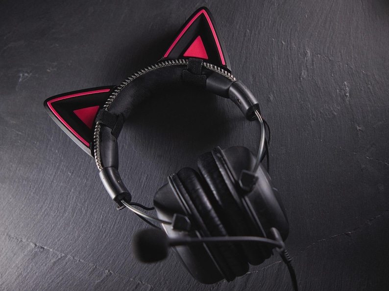 May include: Black headphones with a microphone and pink cat ear covers. The headphones are on a dark grey surface.