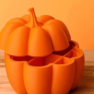 May include: An orange, pumpkin-shaped container with a lid. The container is divided into several compartments. The pumpkin has a stem and is sitting on a wooden surface. The background is a solid orange color.