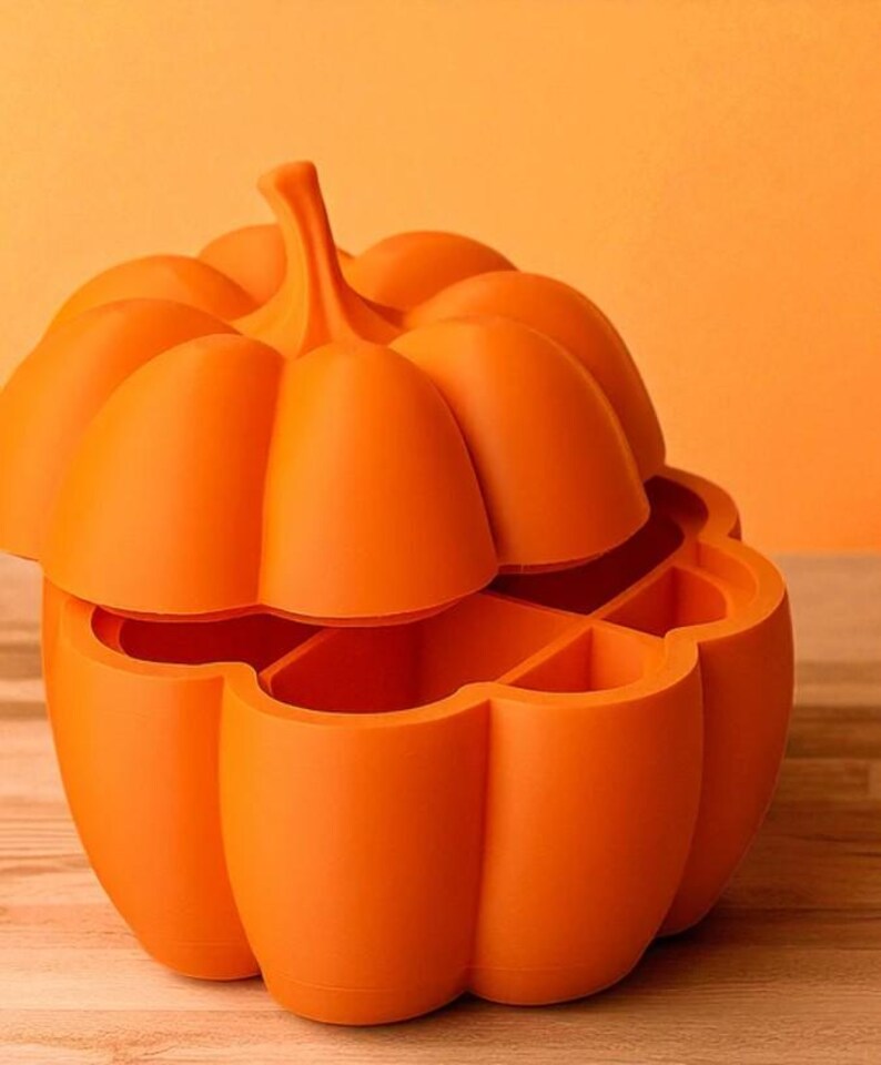 May include: An orange, pumpkin-shaped container with a lid. The container is divided into several compartments. The pumpkin has a stem and is sitting on a wooden surface. The background is a solid orange color.