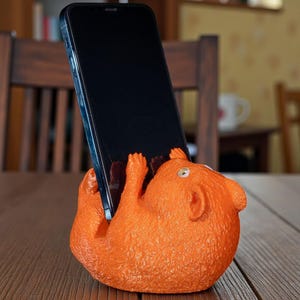 K&ouml;nnte beinhalten: Orangefarbener, eichh&ouml;rnchenf&ouml;rmiger Telefonhalter, der ein schwarzes Smartphone h&auml;lt. Das Eichh&ouml;rnchen hat kleine Pfoten und eine strukturierte Oberfl&auml;che. Der Halter steht auf einem Holztisch.
