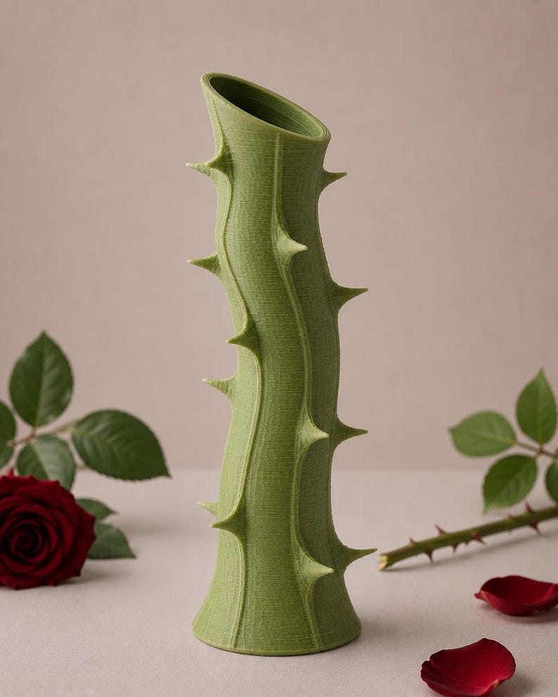May include: A green vase shaped like a thorny stem. The vase has a textured surface and is adorned with several sharp, pointed thorns. The top of the vase is angled. A red rose and petals are in the background.