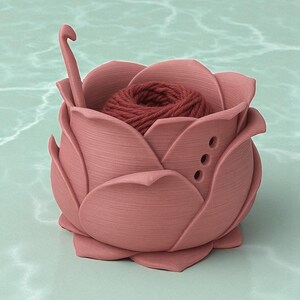 May include: A pink, rose-shaped yarn bowl with a ball of red yarn inside. A crochet hook is placed in the bowl. The bowl has three small holes on the side. The bowl is made of a solid material and sits on a light green surface.