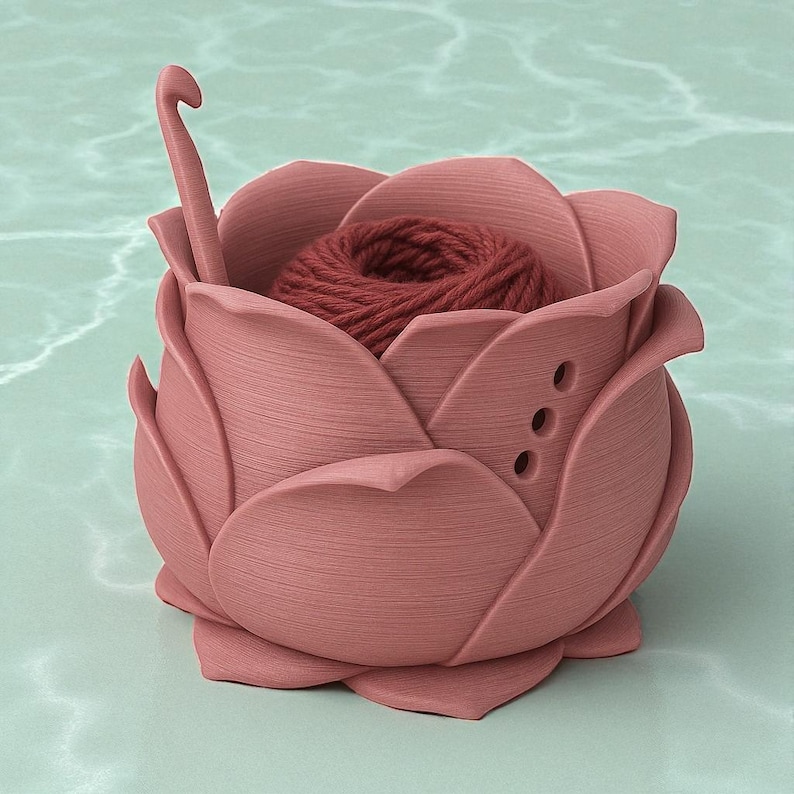 May include: A pink, rose-shaped yarn bowl with a ball of red yarn inside. A crochet hook is placed in the bowl. The bowl has three small holes on the side. The bowl is made of a solid material and sits on a light green surface.